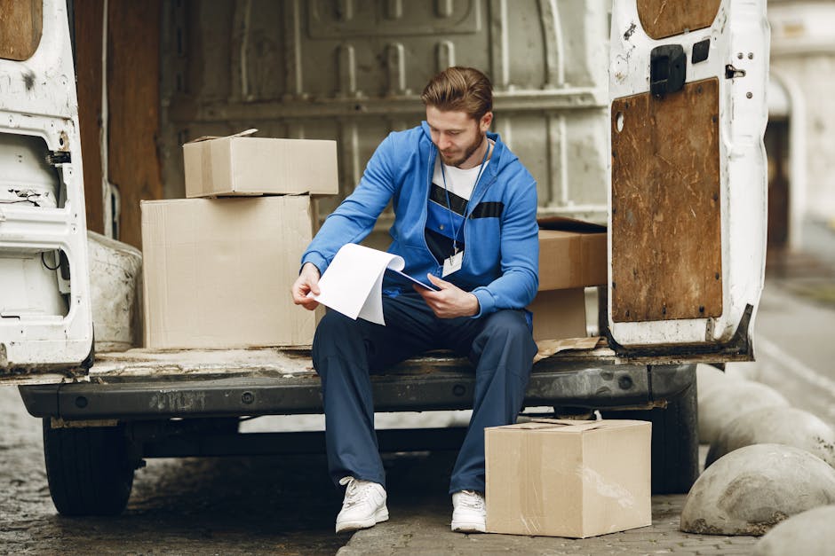 A young male mover with a short, neatly styled haircut is seated on the edge of an open white delivery van in an outdoor area, surrounded by several cardboard boxes of various sizes. He is wearing a s