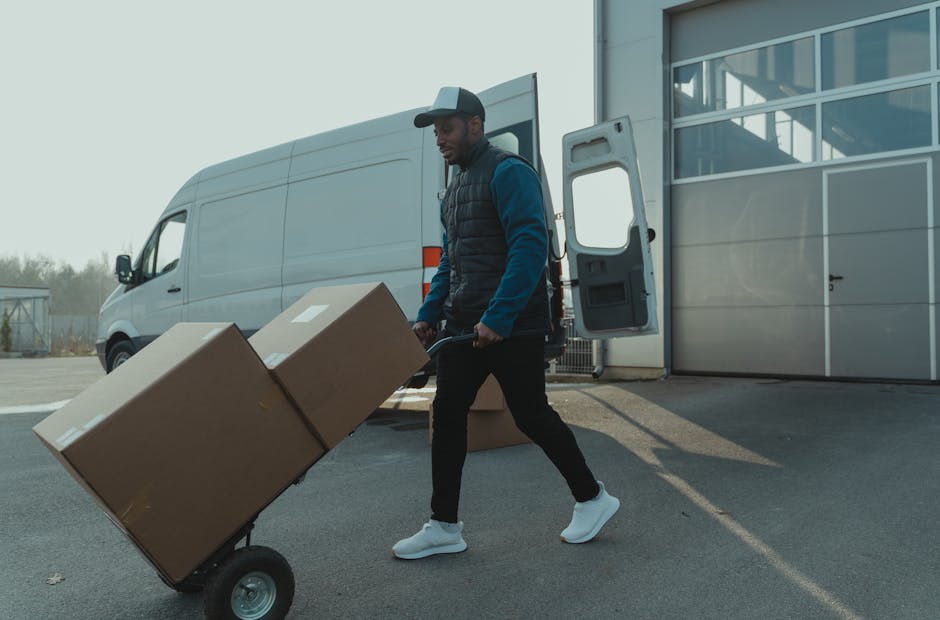 A person wearing a dark jacket, light-colored sneakers, and a cap is loading or unloading a large white van with open rear doors. Inside the van, several brown cardboard boxes of various sizes are sta