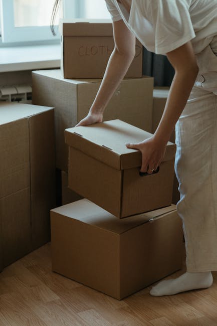 A person wearing a white shirt and light-colored trousers is seen handling a cardboard box during a home relocation process inside a residential property. The individual is in the midst of stacking or