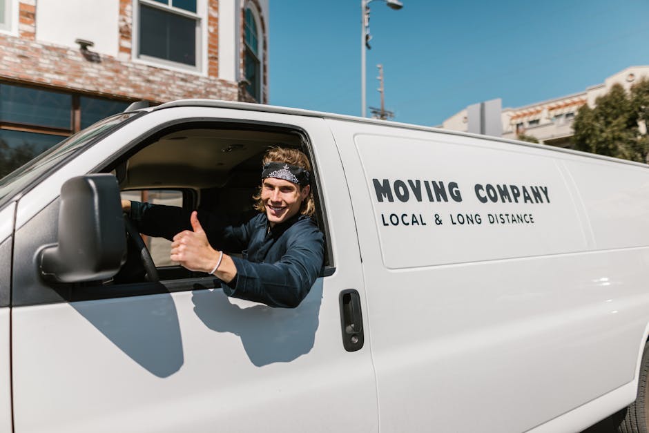 A young male mover with shoulder-length blonde hair, wearing a black bandana and a dark blue jacket, is sitting in the driver's seat of a white moving van parked on a residential street. He is smiling at the camera and giving a thumbs-up gesture through the open window. The van has the words 'MOVING COMPANY LOCAL & LONG DISTANCE' printed on its side. The street background shows modern buildings with brick and glass facades, blue sky, and some greenery. Inside the van, partially visible behind him, are packed cardboard boxes, wrapped furniture, and moving blankets, suggesting preparations for home relocation. The scene captures a moment during a furniture transport or packing and moving process, with the company Man and Van Plumstead providing local moving services, reflecting the logistical aspect of house removals.