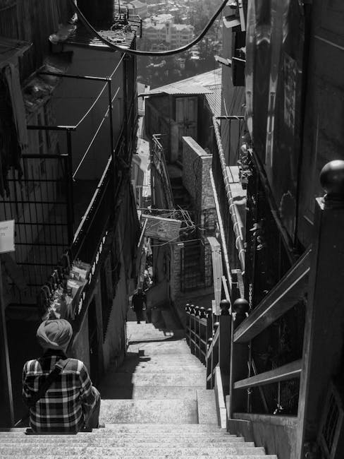 An outside stone staircase with metal railings on both sides ascends between aged white and beige buildings with visible windows and shutters. At the top of the stairs, there is a narrow passage leadi