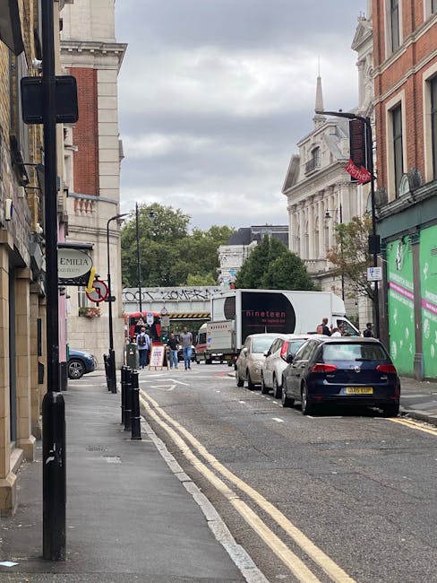 A narrow urban street scene during daytime showing a row of parked cars along the pavement, with a white moving truck labeled 'Dineen' positioned on the road near the curb. Several people are walking 