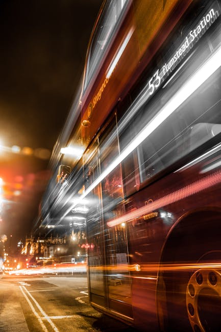Nighttime scene of a red double-decker bus with motion blur, showing the route number 53 and destination Plumstead Station, traveling along a city street illuminated by streetlights and vehicle headlights. The bus is adjacent to a curb with visible road markings, and the background includes blurred cityscape elements such as buildings and traffic. This image captures the movement associated with urban transport, relevant to relocation logistics and moving services provided by Man and Van Plumstead, especially for clients planning home relocation or furniture transport in the Plumstead Common to Woolwich Arsenal area.
