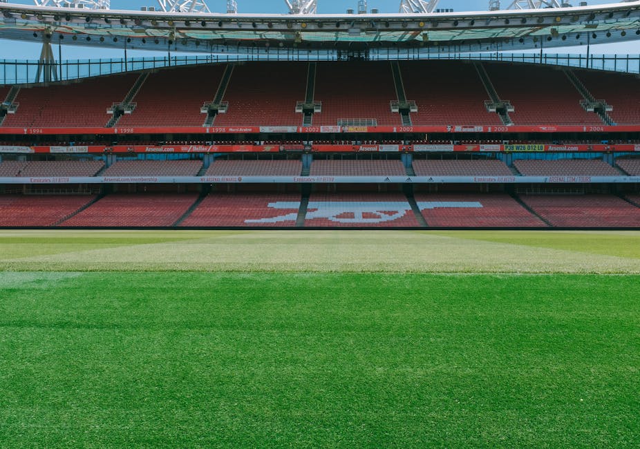 An empty football stadium with several tiers of seating painted in red, with some banners displaying the Arsenal football club name and logo, visible in the background. The playing field features well-maintained grass with varying shades of green, divided into different sections, and is illuminated by natural daylight. The stadium includes a large, curved roof structure supported by visible beams and trusses, covering parts of the seating area. The scene is devoid of spectators or players, focusing on the clean, organized environment suitable for a home relocation process involving planning or viewing before an event, as would be relevant to a professional removals and moving service like Man and Van Plumstead.