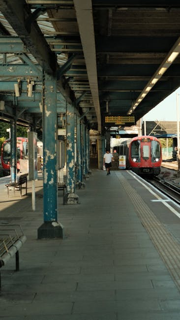 A view of a railway station platform during daytime with two individuals walking along the paved walkway beside the train tracks. The platform is equipped with signage, including platform numbers 3 an
